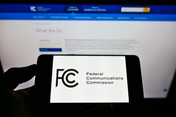 Person holding mobile phone with seal of US agency Federal Communications Commission (FCC) on screen in front of web page. Focus on phone display