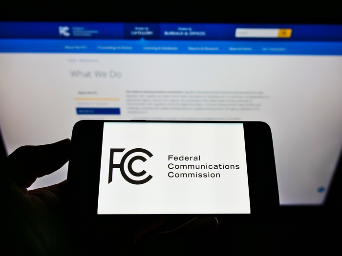 Person holding mobile phone with seal of US agency Federal Communications Commission (FCC) on screen in front of web page. Focus on phone display