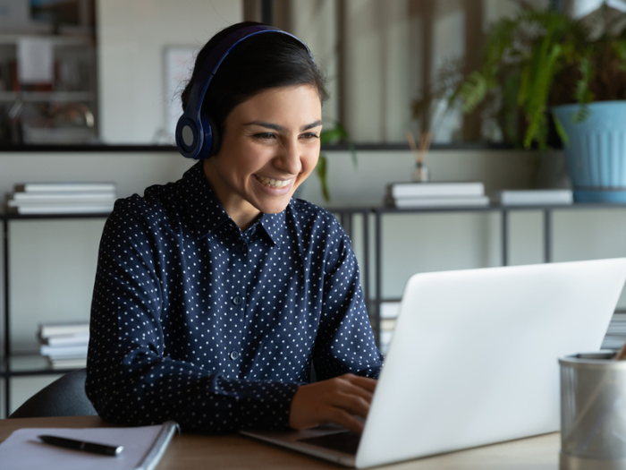 Head shot happy young indian ethnicity female manager wearing wireless headphones, looking at laptop screen, holding pleasant conversation with partners clients online, working remotely at workplace.