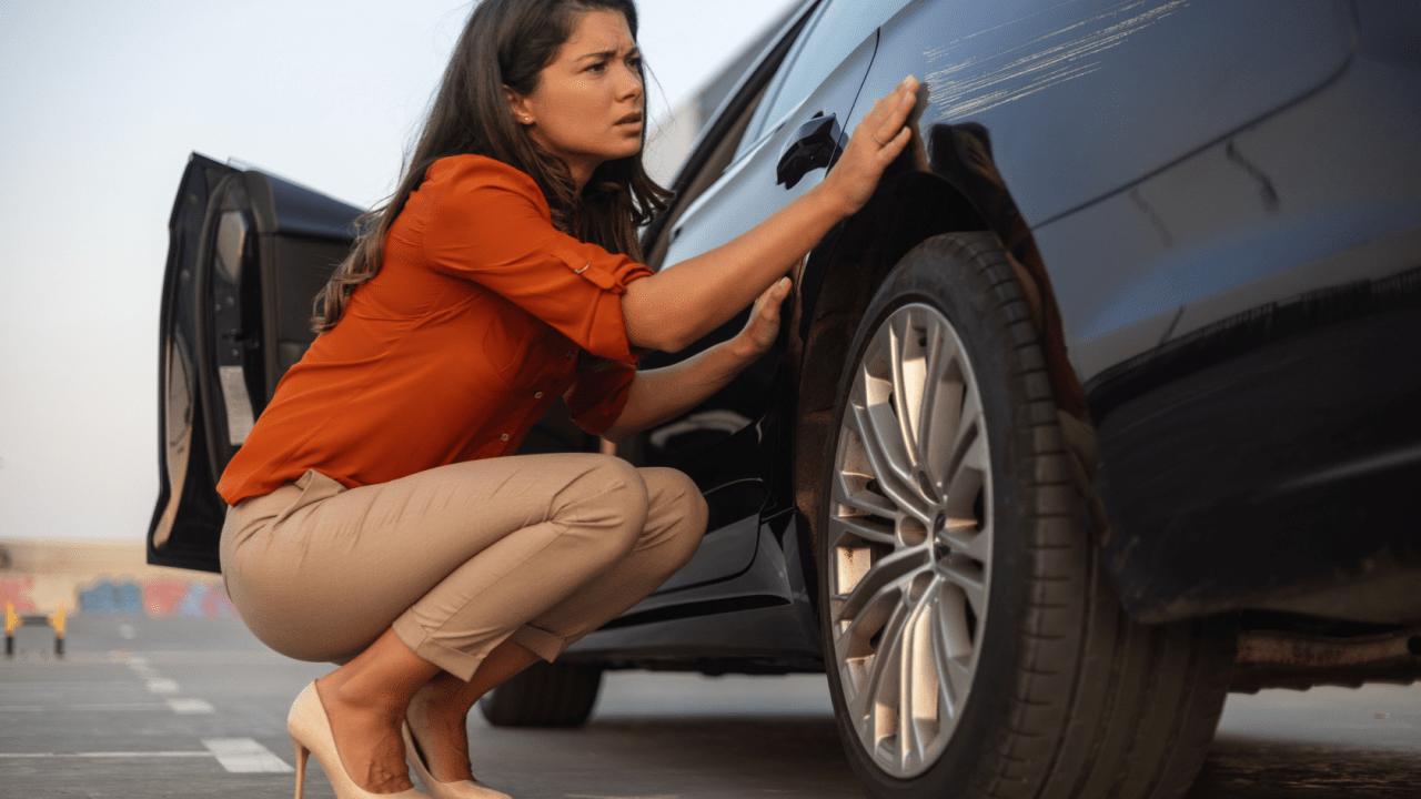 Woman in orange shirt examining car damage while crouched next to vehicle, considering whether it's better to pay out of pocket for a car accident