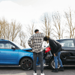 Two person examining rear-end collision damage between blue and black cars, showing what happens if someone wrecks your car and they aren't on your insurance Progressive
