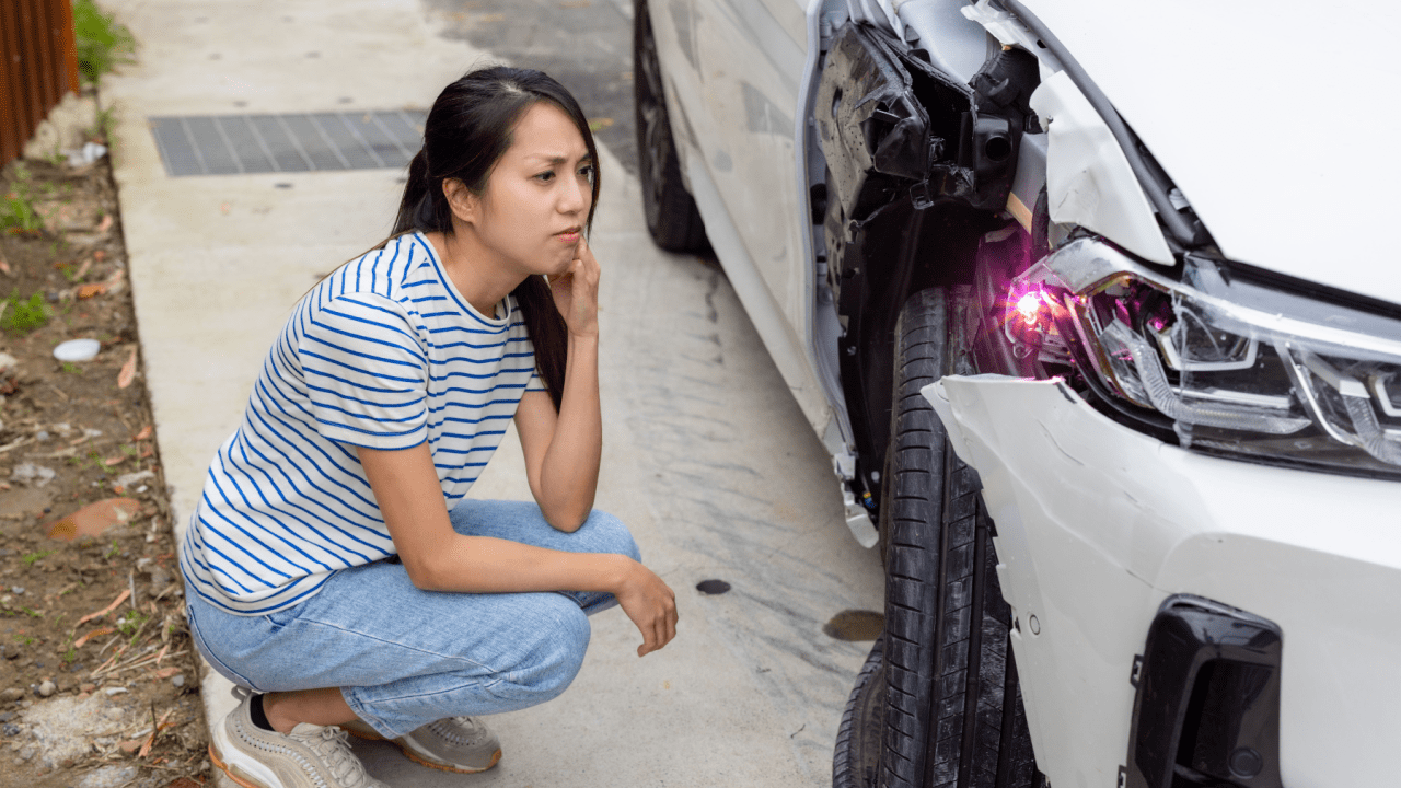 Woman in striped shirt looking distressed while crouching next to damaged white car, illustrating what happens if you cause a car accident but don't hit anyone
