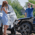 Woman on phone and stressed man examining collision damage between white SUV and dark car, demonstrating what is considered an at-fault accident