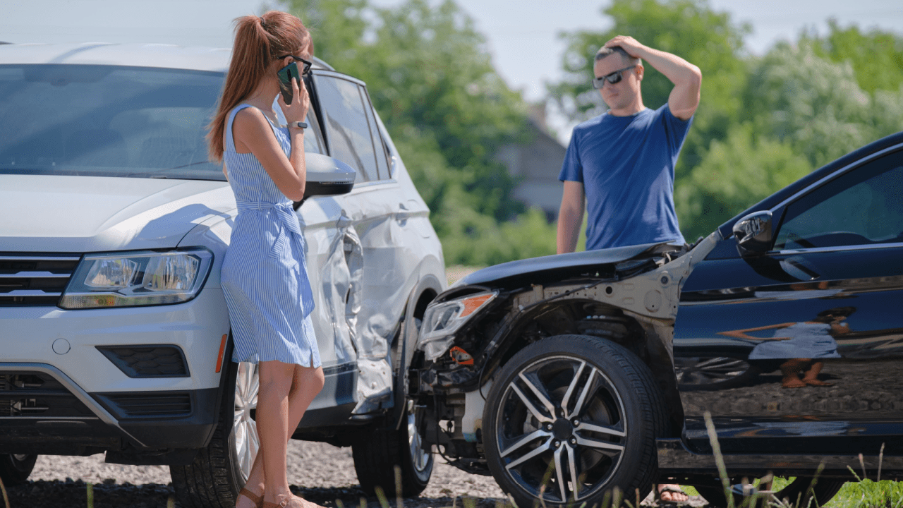 Woman on phone and stressed man examining collision damage between white SUV and dark car, demonstrating what is considered an at-fault accident