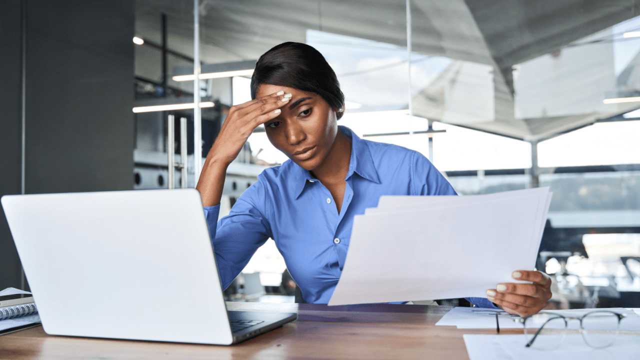Stressed businesswoman in blue shirt holding head while reviewing financial documents at laptop, discovering what is the number one cost of bankruptcy