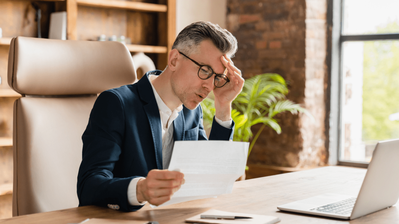 Stressed businessman in suit and glasses holding bankruptcy documents while working at laptop, showing signs of financial distress