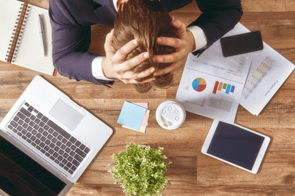 Overhead view of stressed businessman with hands on head surrounded by financial documents, laptop, and tablet while researching corporate bankruptcy filings