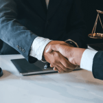 Two legal professionals shaking hands across a desk with a gavel, laptop, and justice scale — how to get new legal clients.
